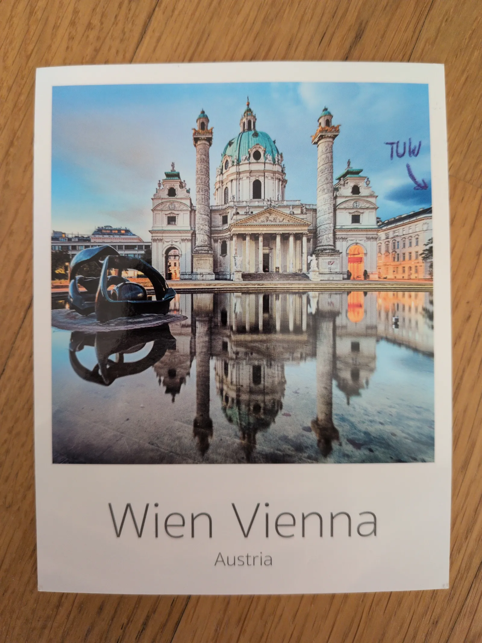 Ein Postkartenbild der Wiener Karlskirche mit einer Skulptur im Vordergrund, die sich beide im Wasser spiegeln, und dem Text "Wien Vienna Austria".