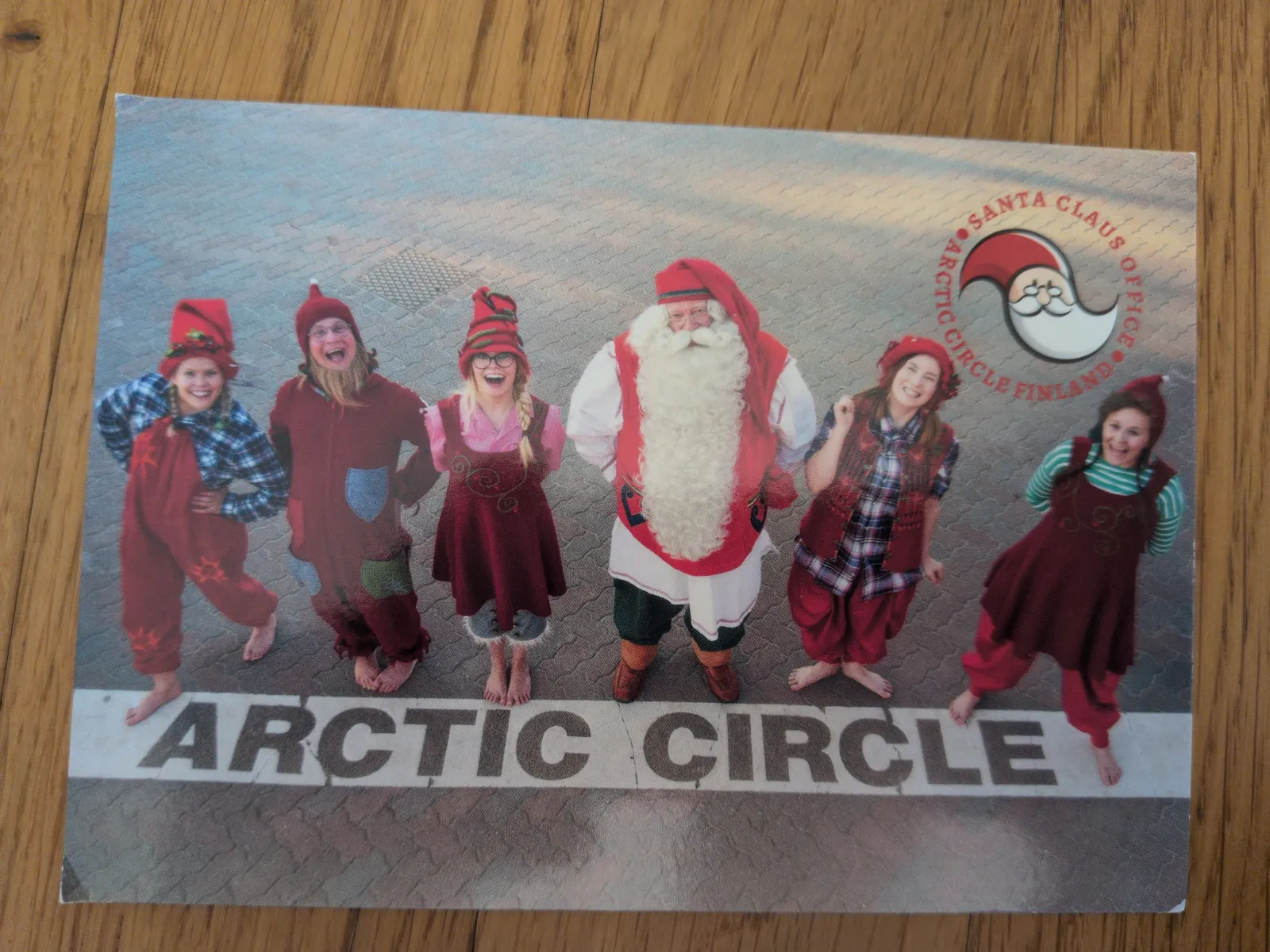 Der Weihnachtsmann und fünf lachende Elfen stehen barfuß auf einem gepflasterten Boden, auf dem "ARCTIC CIRCLE" geschrieben steht.
