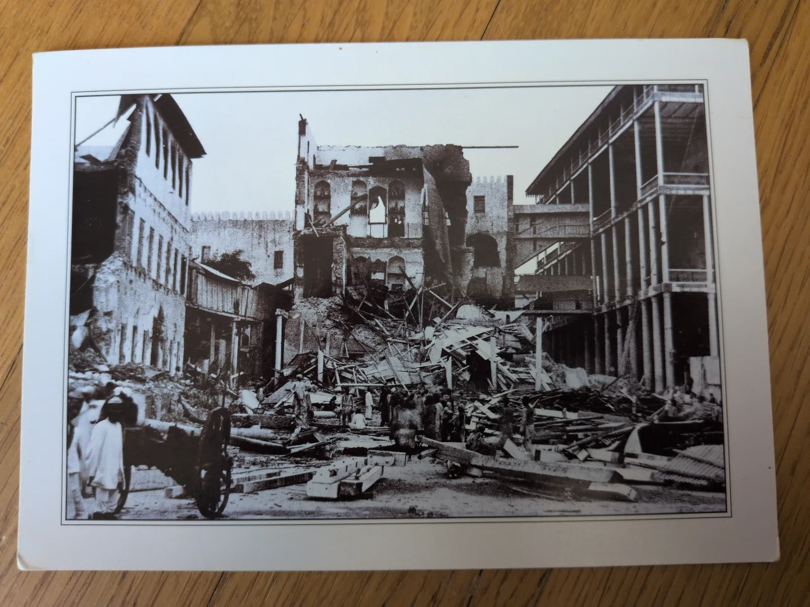 Ein Schwarz-Weiß-Foto zeigt eine zerstörte Stadtstraße voller Schutt und Trümmer, mit teils eingestürzten Gebäuden und einigen Menschen, die sich darin bewegen.
