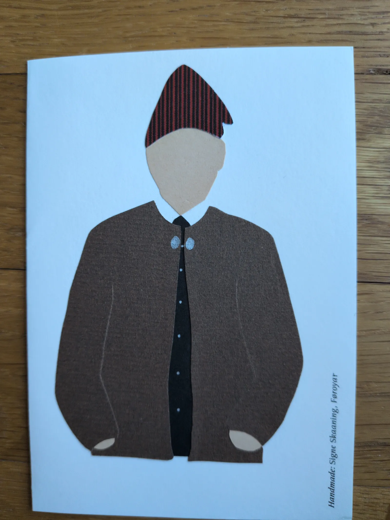Ein Papierschnitt zeigt eine männliche Figur in brauner Tracht mit einem rot-schwarz gestreiften Hut und silbernen Spangen auf hellblauem Grund.