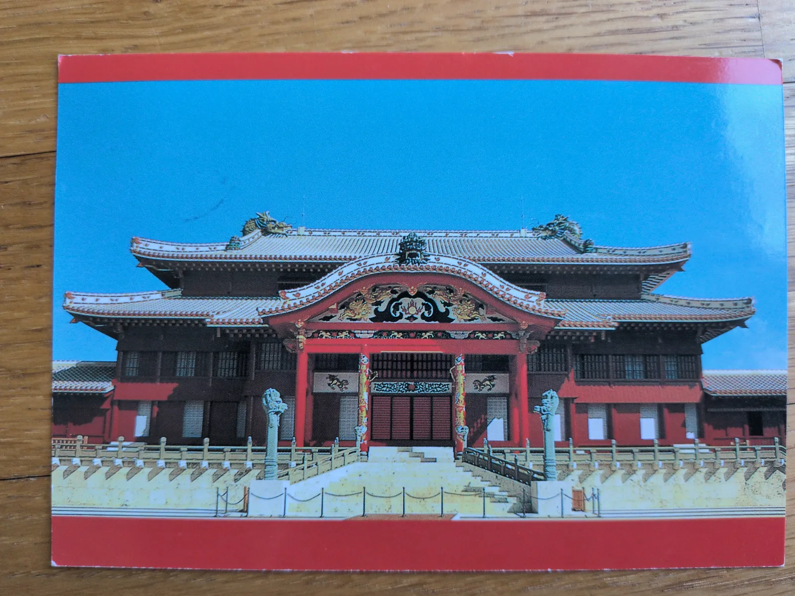 Ein rotes, mehrstöckiges traditionelles Palastgebäude mit aufwendig verziertem Eingang steht unter blauem Himmel, gerahmt von einem roten Rand.