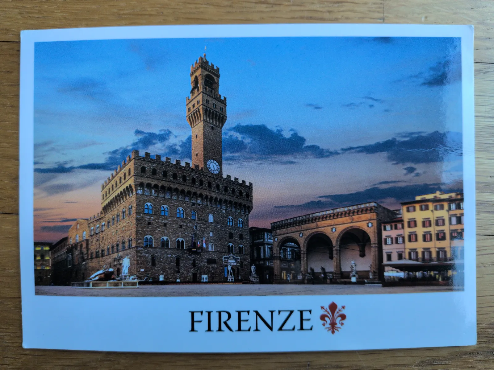 Ein Postkartenmotiv von Florenz zeigt den Palazzo Vecchio mit seinem hohen Turm und der Loggia dei Lanzi bei Dämmerung.
