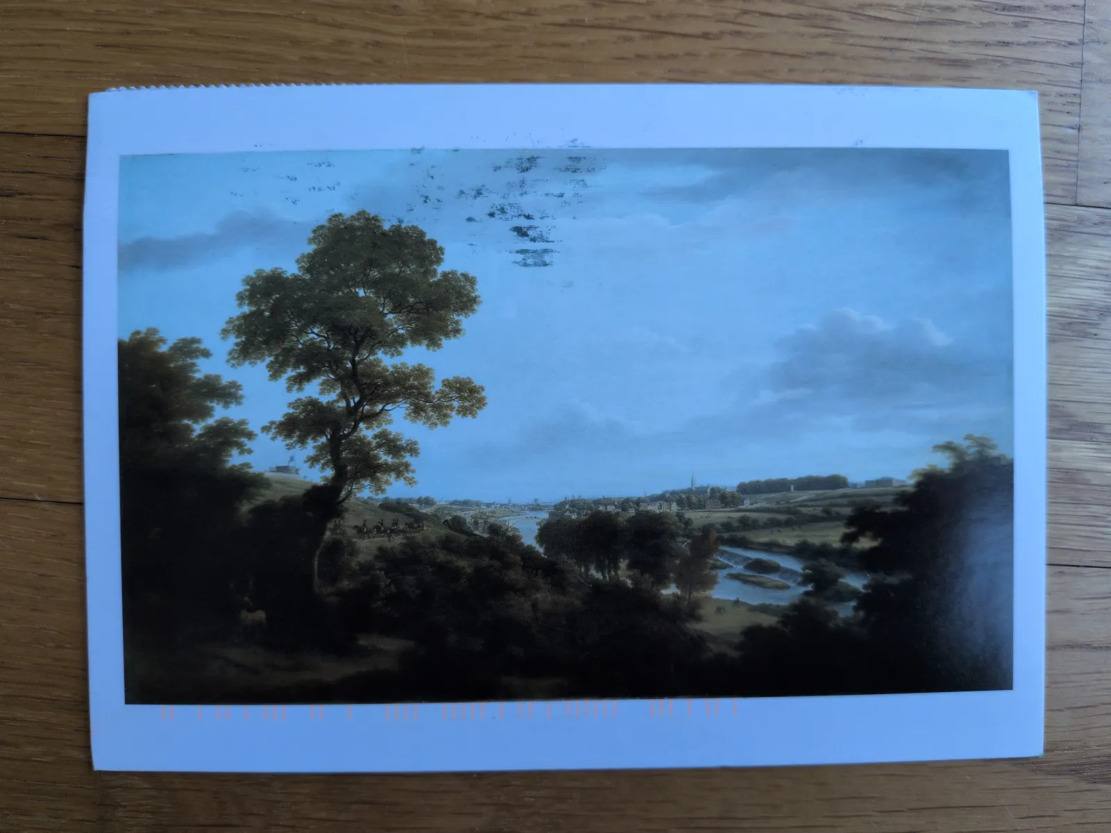 Ein klassisches Landschaftsgemälde zeigt eine weite Flusslandschaft mit einer Stadt, bewaldeten Hügeln und einem großen Baum unter blauem Himmel.
