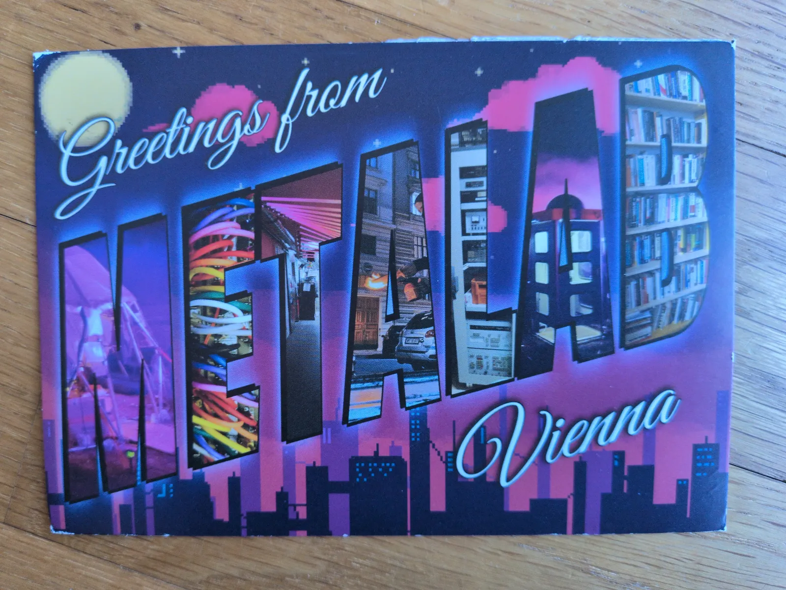 Eine Postkarte zeigt den Schriftzug "Greetings from METALAB Vienna", dessen Großbuchstaben aus farbigen Collagen von Stadtansichten und Innenräumen bestehen, vor einem stilisierten Nachthimmel mit pinken Wolken und dunkler Skyline.