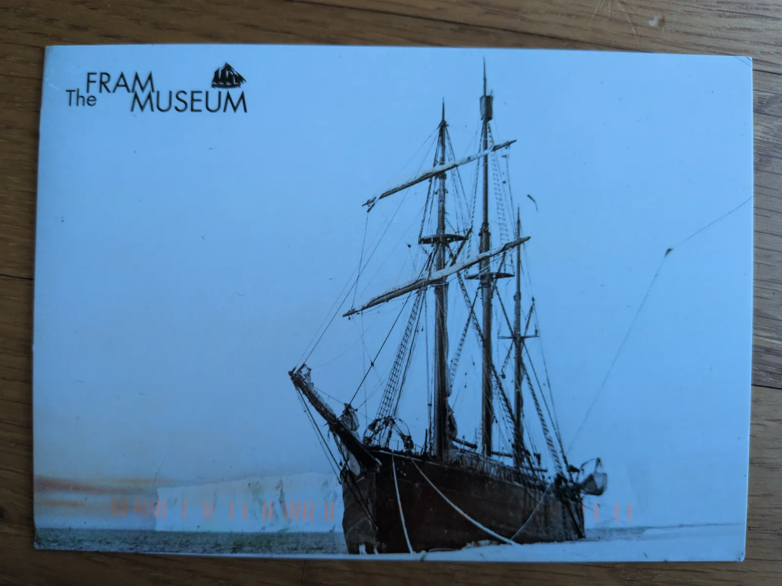 Ein historisches dreimastiges Segelschiff steckt im Eis vor einer hellen Eiswand, mit dem Logo des Fram Museums oben links.