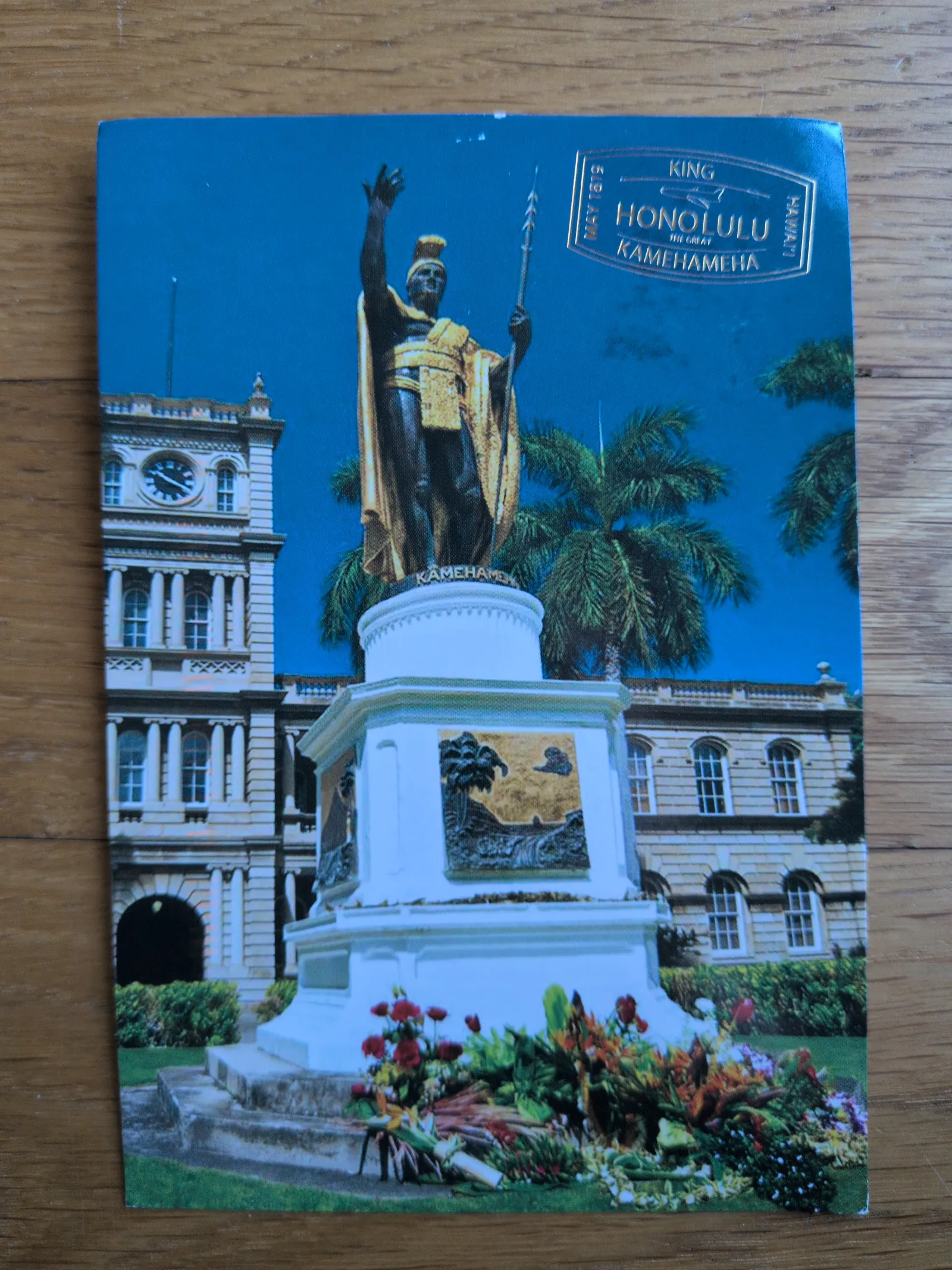 Eine goldene Statue von König Kamehameha steht unter blauem Himmel vor einem historischen Gebäude mit Uhrturm, umgeben von Palmen und Blumen.