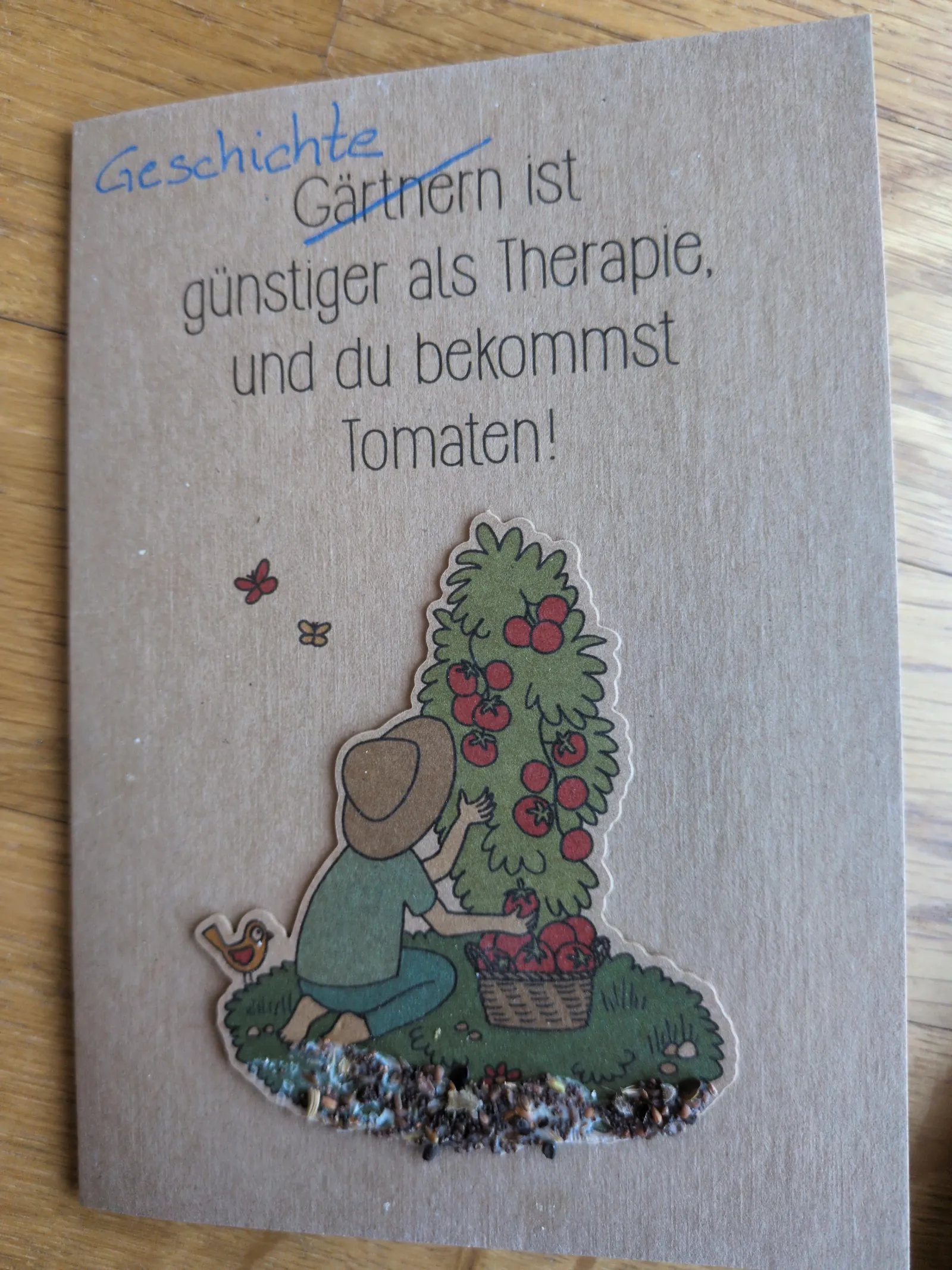 Eine braune Karte zeigt den Spruch "Gärtnern ist günstiger als Therapie, und du bekommst Tomaten!",  hier ist das Wort "Gärtnern" durchgestrichen und  "Geschichte" darüber geschrieben. Darunter eine Illustration einer Person, die Tomaten erntet, und aufgeklebte Samen am unteren Rand.