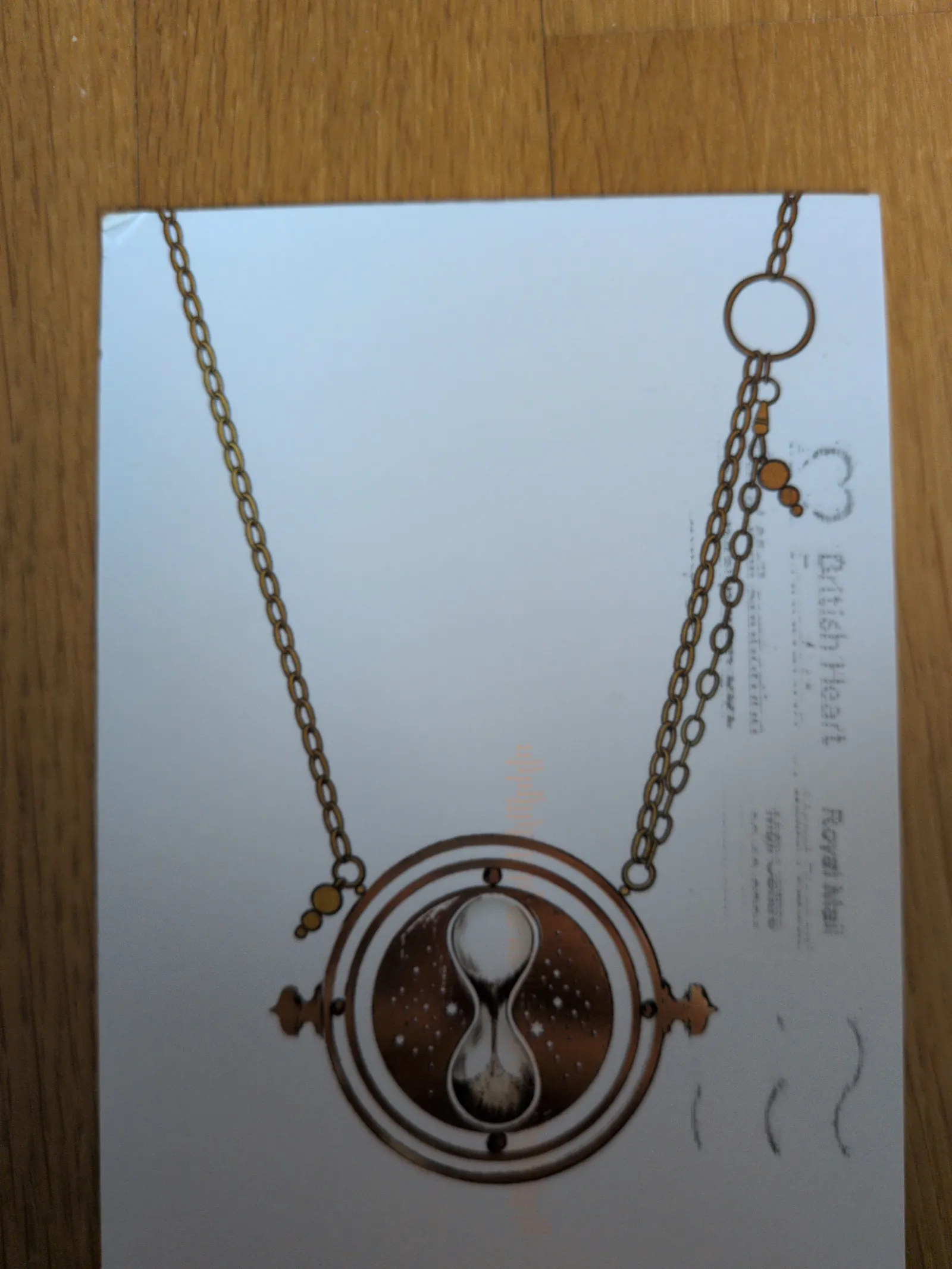 Das Bild zeigt eine goldene Kette mit einem Sanduhr-Anhänger auf weißem Grund, daneben schwache Stempelabdrücke.