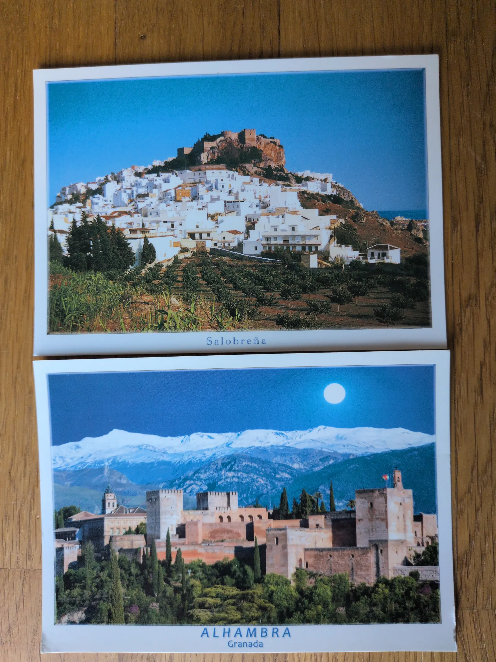 Zwei Postkarten liegen auf einem Holztisch: Die obere zeigt Salobreña, ein weißes Dorf mit Burg auf einem Hügel, die untere die Alhambra in Granada vor schneebedeckten Bergen bei Mondschein.