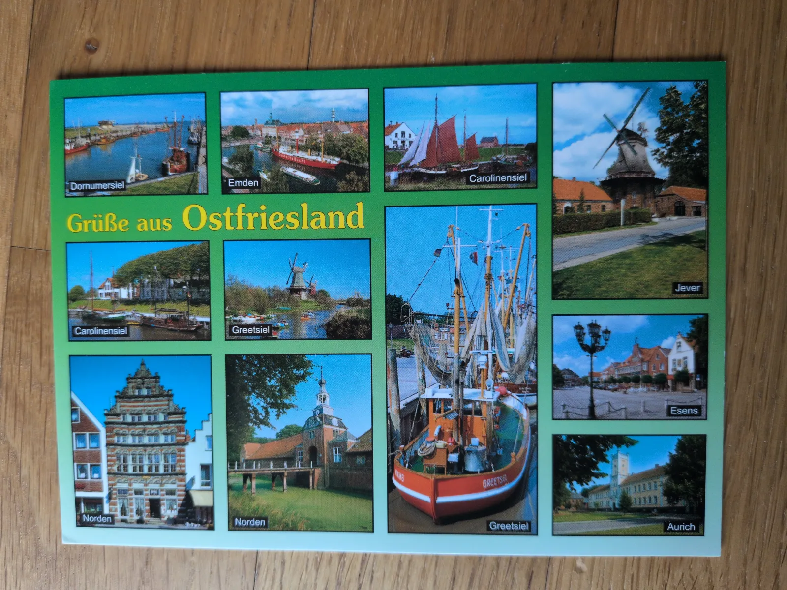 Die Postkarte zeigt eine Collage aus zwölf Bildern von Ostfriesland, darunter Häfen mit Booten, Windmühlen und historische Gebäude.