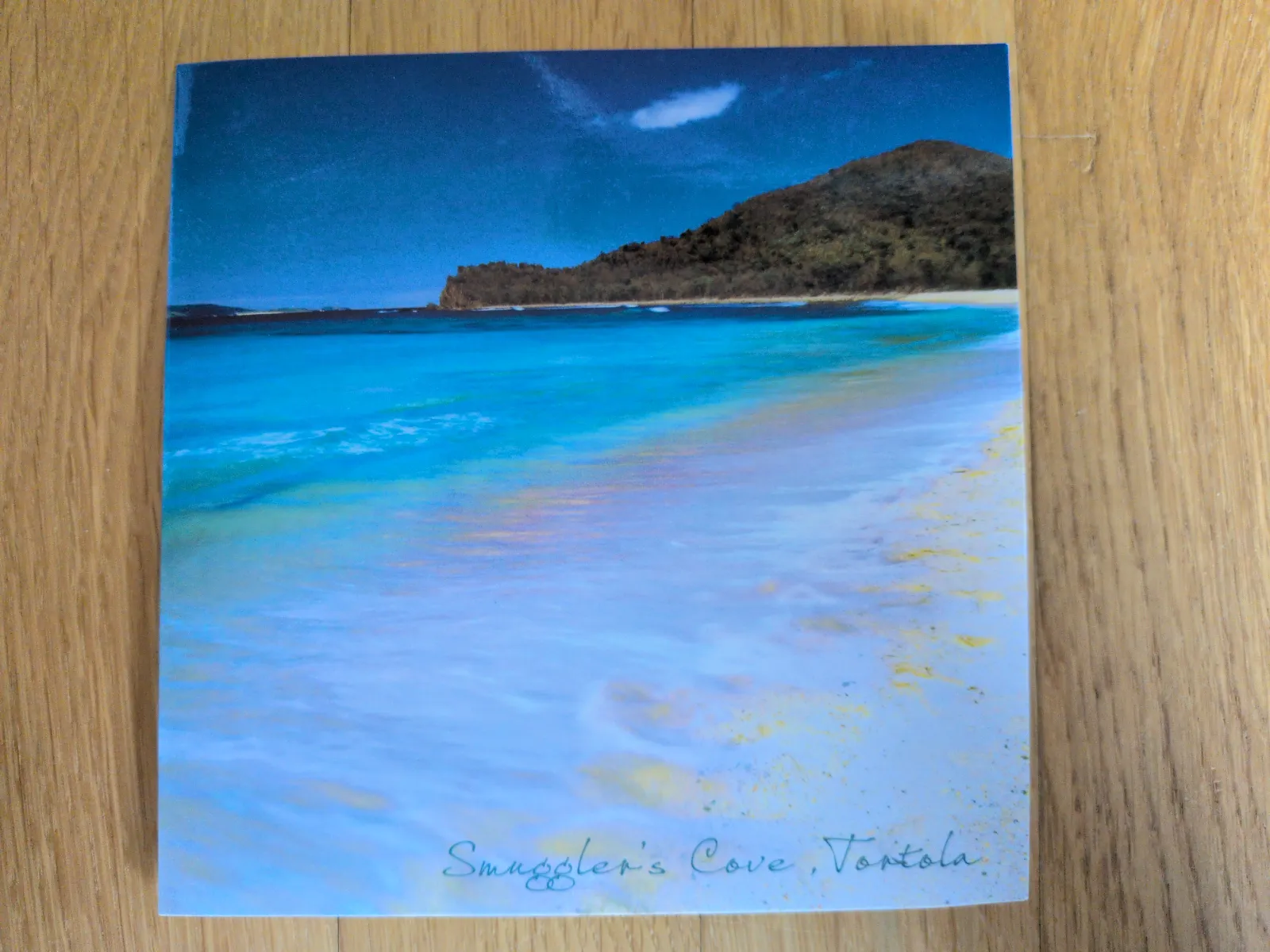 Das Bild zeigt einen tropischen Strand von Smuggler's Cove, Tortola, mit türkisfarbenem Meer, das sanft an den hellen Sand spült, und grünen Hügeln unter einem blauen Himmel.