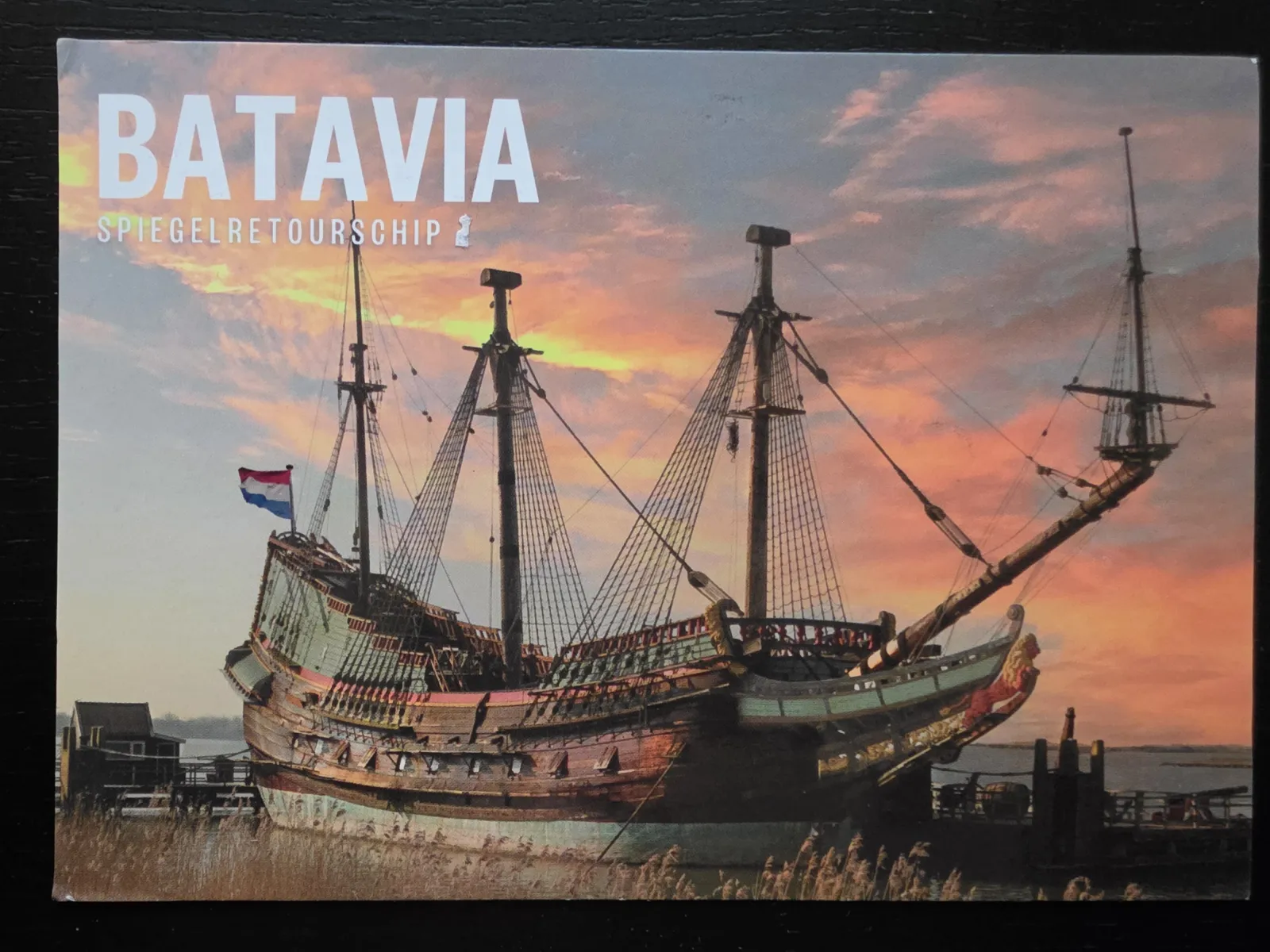 Ein großes, historisches Segelschiff mit drei Masten und Takelage liegt am Ufer vor einem farbenprächtigen Sonnenuntergangshimmel; oben links im Bild steht der Schriftzug „BATAVIA“.