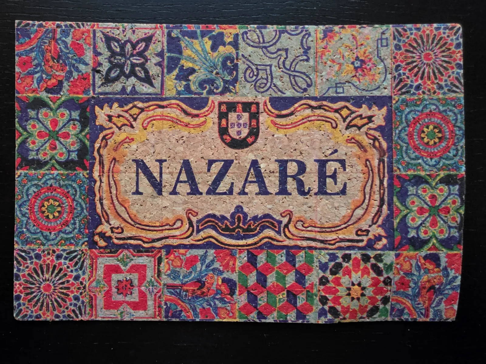 Ein bunter Kork-Untersetzer zeigt in der Mitte den Schriftzug "NAZARÉ" und ein Wappen, umrahmt von einem Mosaik aus vielfältigen, farbenfrohen portugiesischen Fliesenmustern.