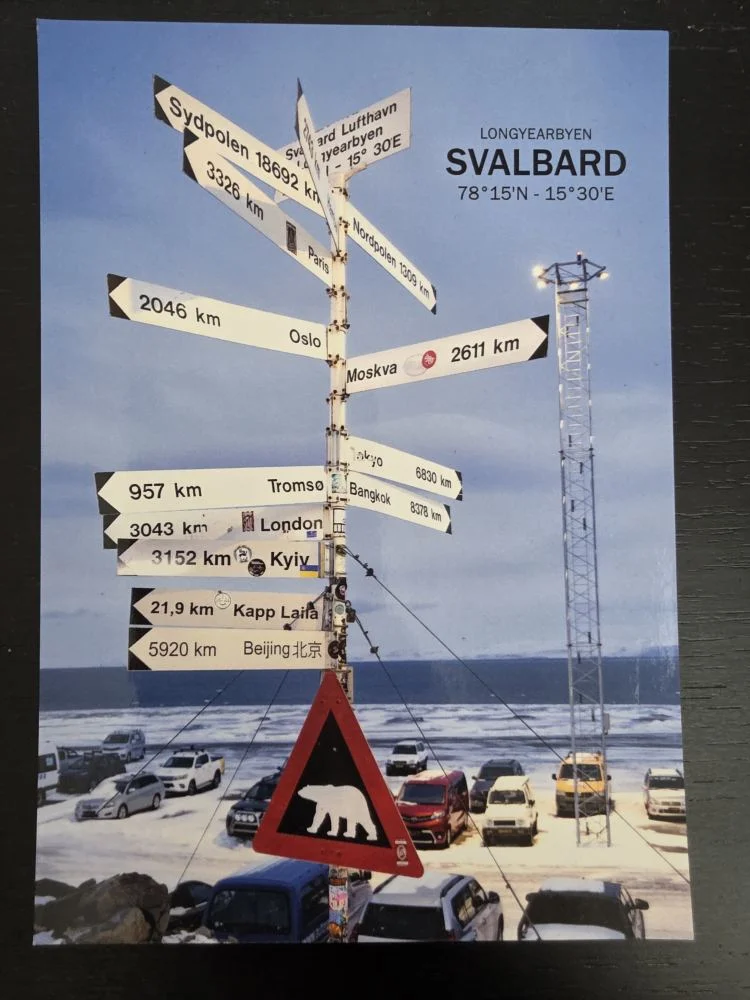 Ein Wegweiser auf Svalbard zeigt Entfernungen zu Städten weltweit, darunter ein Eisbären-Warnschild und zahlreiche geparkte Autos in einer winterlichen Landschaft.
