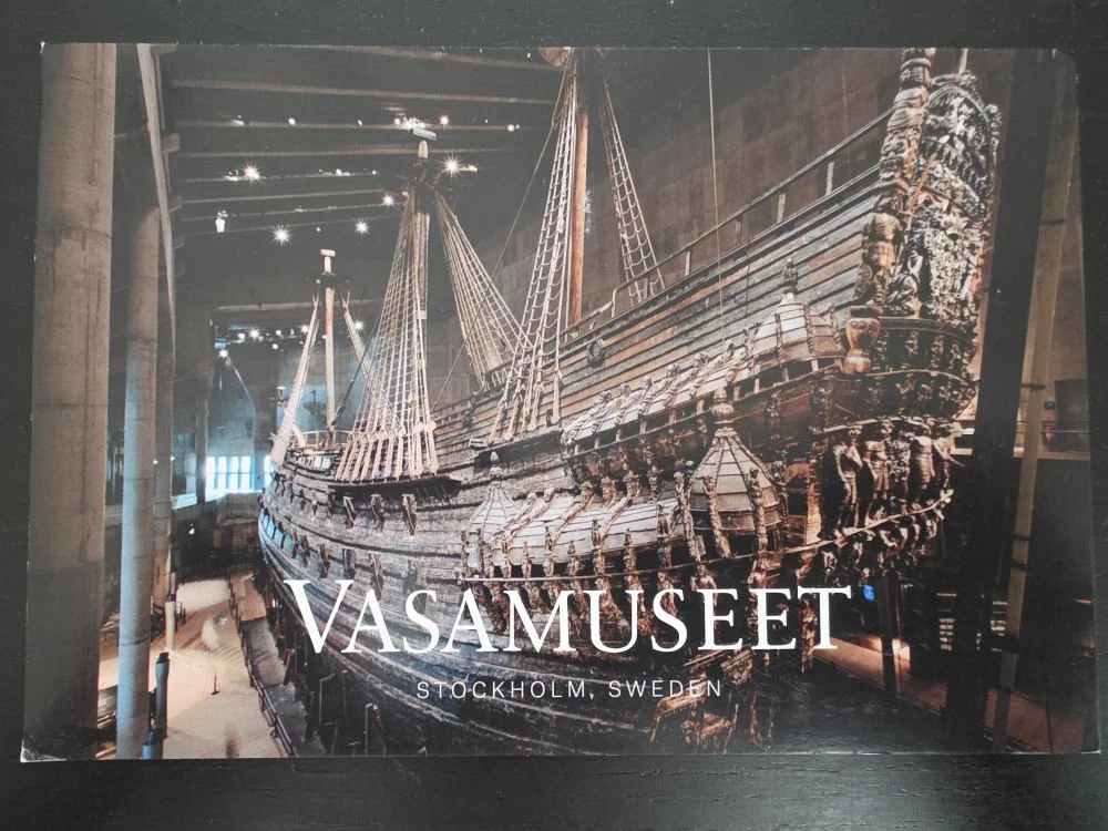 Ein riesiges, reich verziertes Holzschiff mit Masten und Takelage ist in einem dunklen Museum ausgestellt, mit dem Schriftzug „Vasamuseet“ im Vordergrund.
