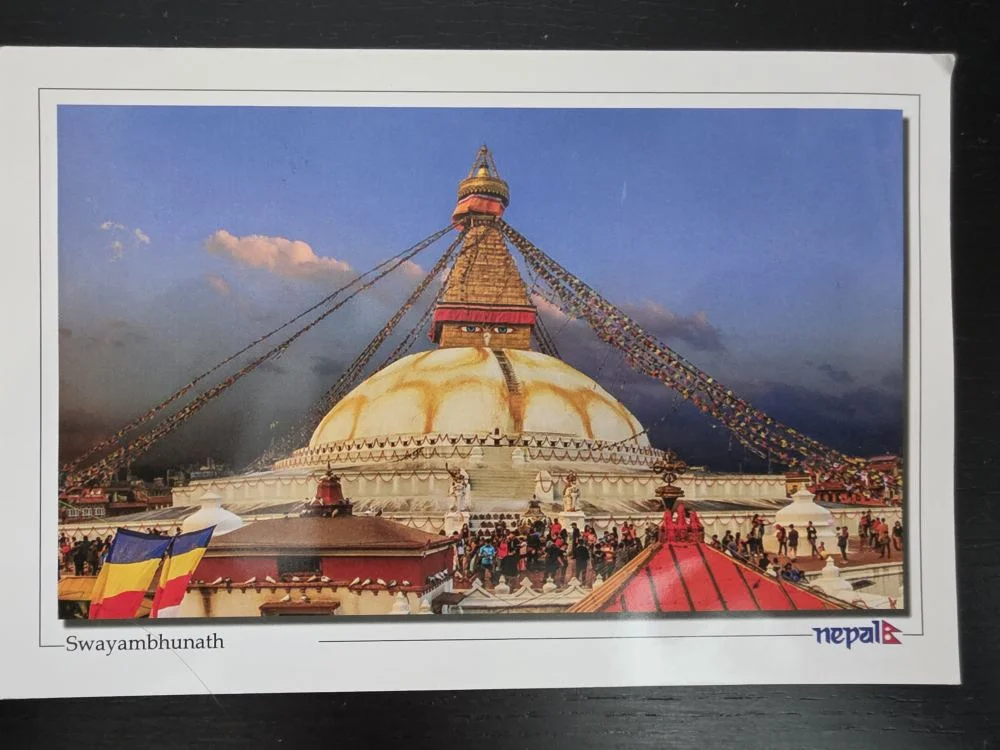 Ein großes, weißes buddhistisches Stupa mit einer goldenen Spitze, auf der die Augen Buddhas gemalt sind, und bunten Gebetsfahnen, umgeben von Menschen.