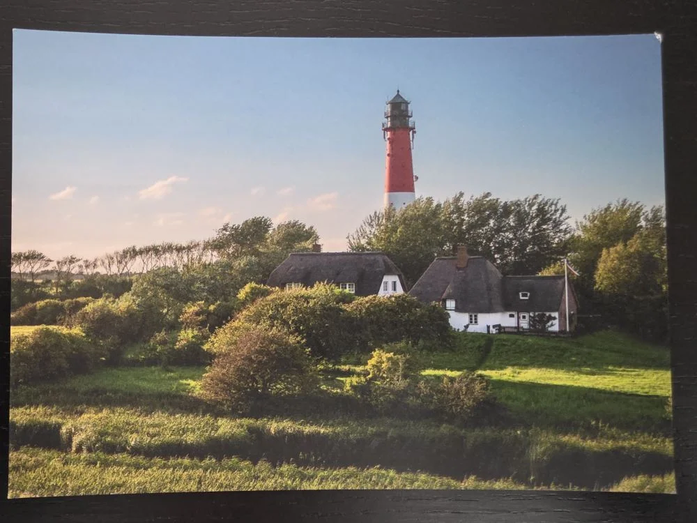 Ein roter und weißer Leuchtturm ragt über zwei reetgedeckte Häuser in einer weiten grünen Landschaft.
