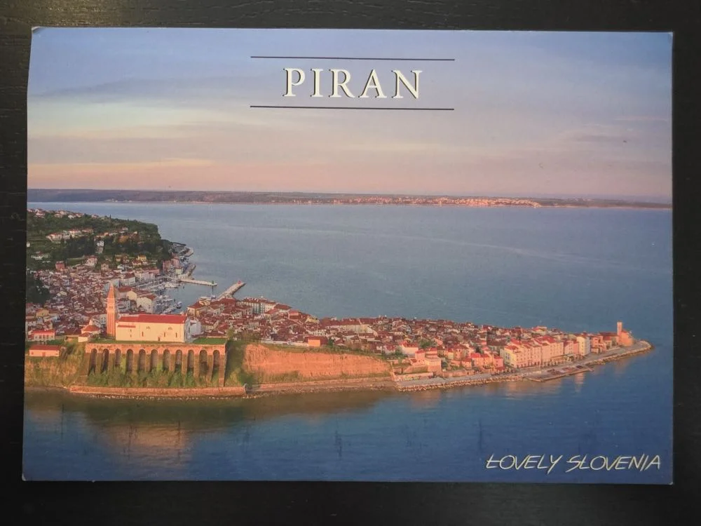 Eine Postkarte zeigt eine Luftaufnahme der malerischen Halbinsel von Piran in Slowenien im warmen Abendlicht, umgeben von blauem Meer.