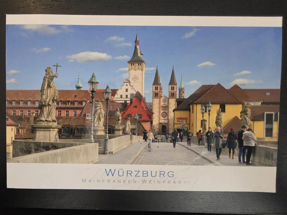 Eine Postkarte zeigt die Alte Mainbrücke in Würzburg mit ihren steinernen Heiligenstatuen und Fußgängern vor der Kulisse historischer Türme unter blauem Himmel.