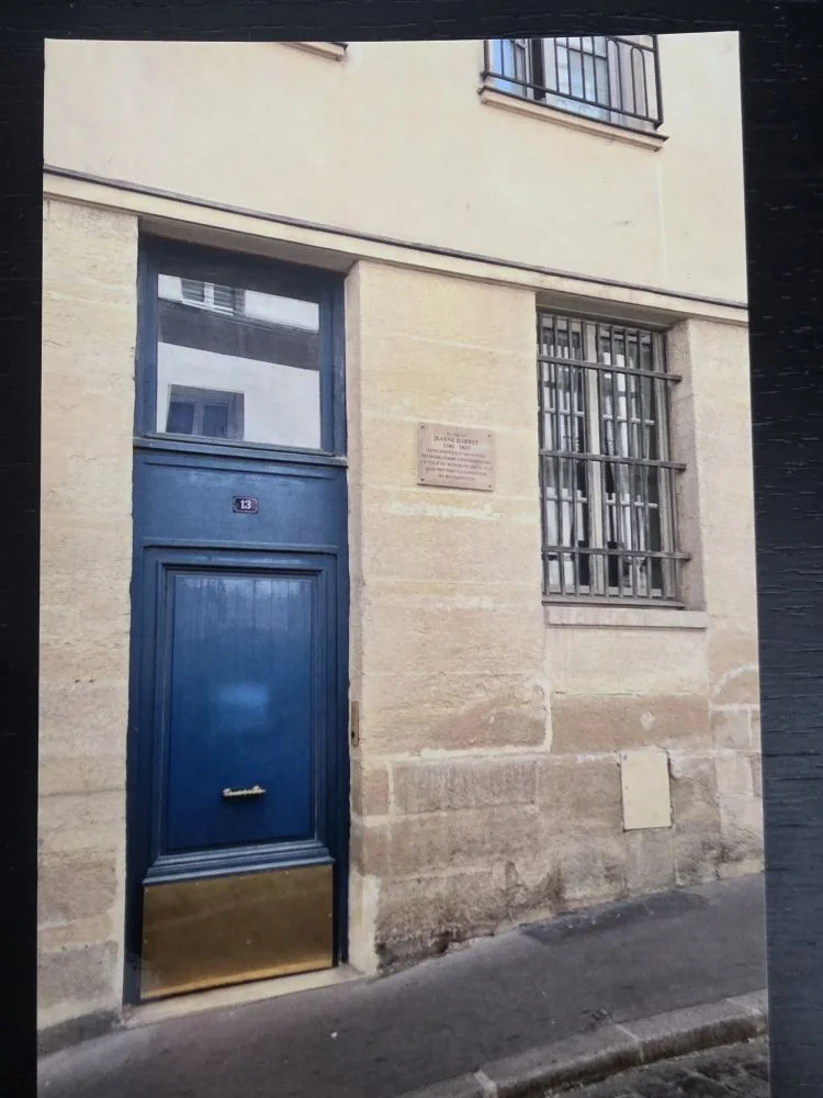 Eine beige Sandsteinfassade zeigt eine dunkelblaue Eingangstür mit der Nummer 13, neben der sich eine kleine Gedenktafel und ein vergittertes Fenster befinden.