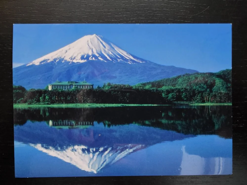 Der schneebedeckte Fuji spiegelt sich zusammen mit einem bewaldeten Ufer und einem Gebäude perfekt in ruhigem, blauen Wasser.