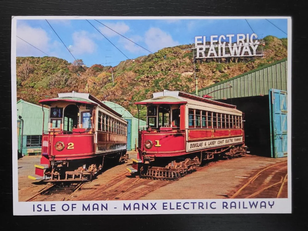 Zwei historische, rot-beige Straßenbahnwagen mit den Nummern 1 und 2 stehen nebeneinander auf Schienen vor einem Depot der Manx Electric Railway.