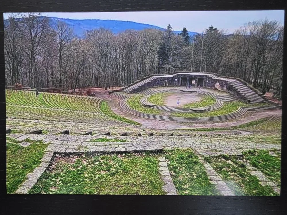Ein großes, von dichtem Wald umgebenes Freilicht-Amphitheater aus Stein mit halbkreisförmigen, grasbewachsenen Zuschauerrängen.