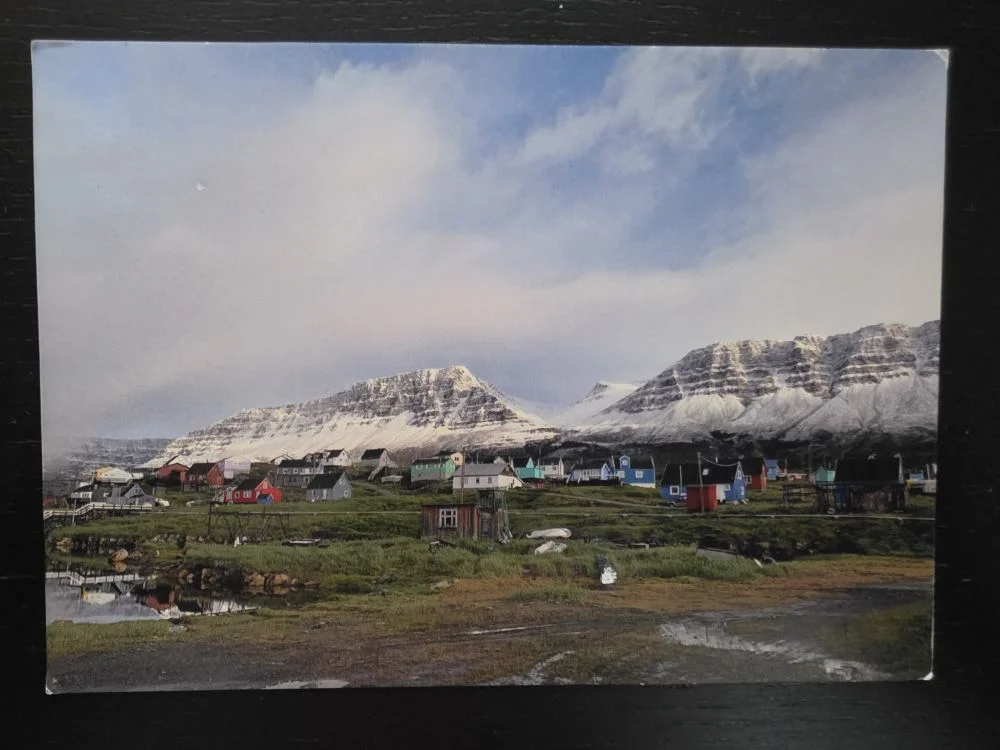 Ein kleines Dorf mit bunten Holzhäusern liegt auf einer grünen Wiese vor einer Kulisse aus majestätischen, schneebedeckten Bergen unter einem bewölkten Himmel.