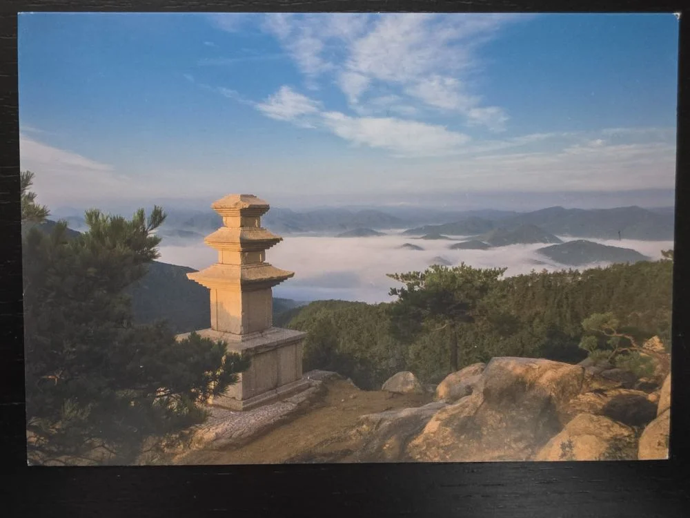 Eine steinerne Pagode steht auf einem felsigen Berggipfel über einem weiten Nebelmeer, aus dem ferne Berggipfel unter blauem Himmel herausragen.
