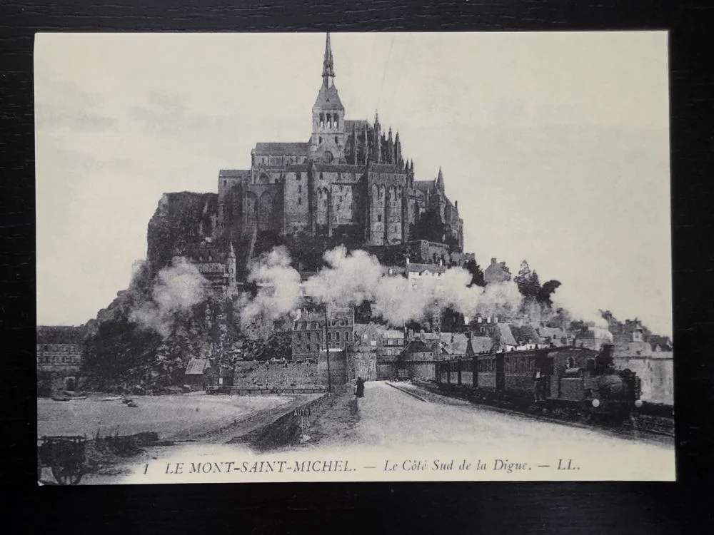 Eine historische Schwarz-Weiß-Postkarte zeigt die Klosterinsel Mont-Saint-Michel mit einer dampfenden Eisenbahn auf dem Deich im Vordergrund.