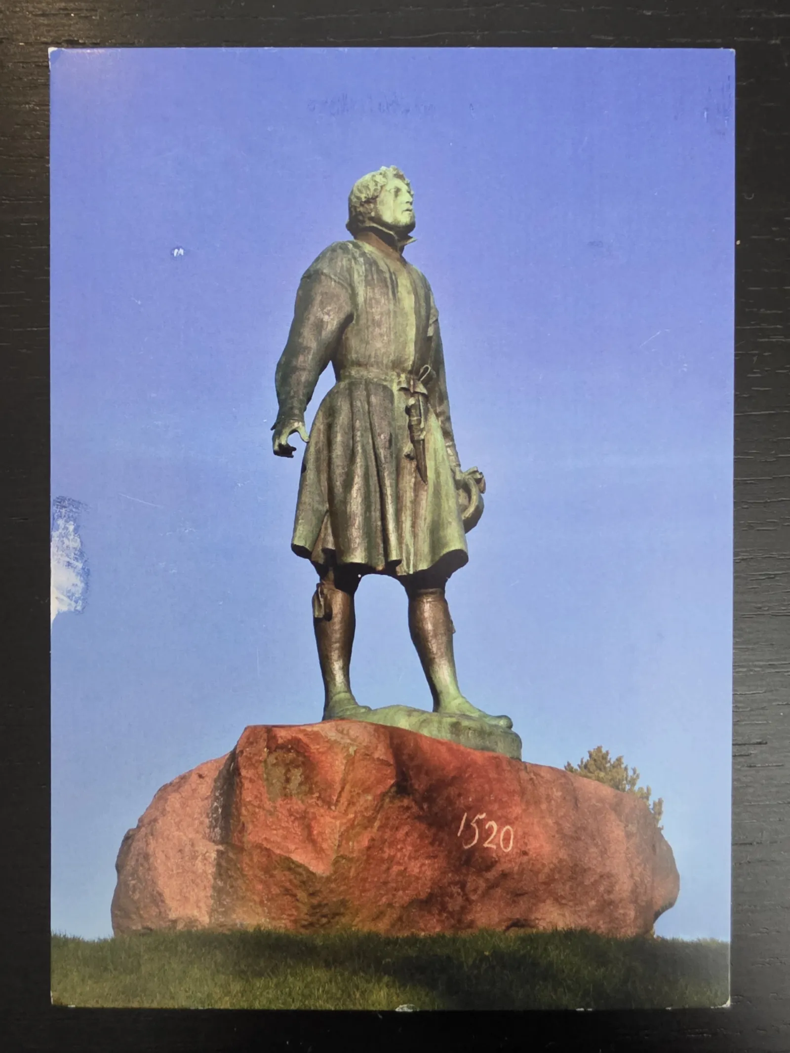 Eine Bronzestatue eines Mannes in historischer Kleidung steht auf einem großen, rötlichen Felsen mit der Jahreszahl „1520“ vor einem klaren, blauen Himmel.