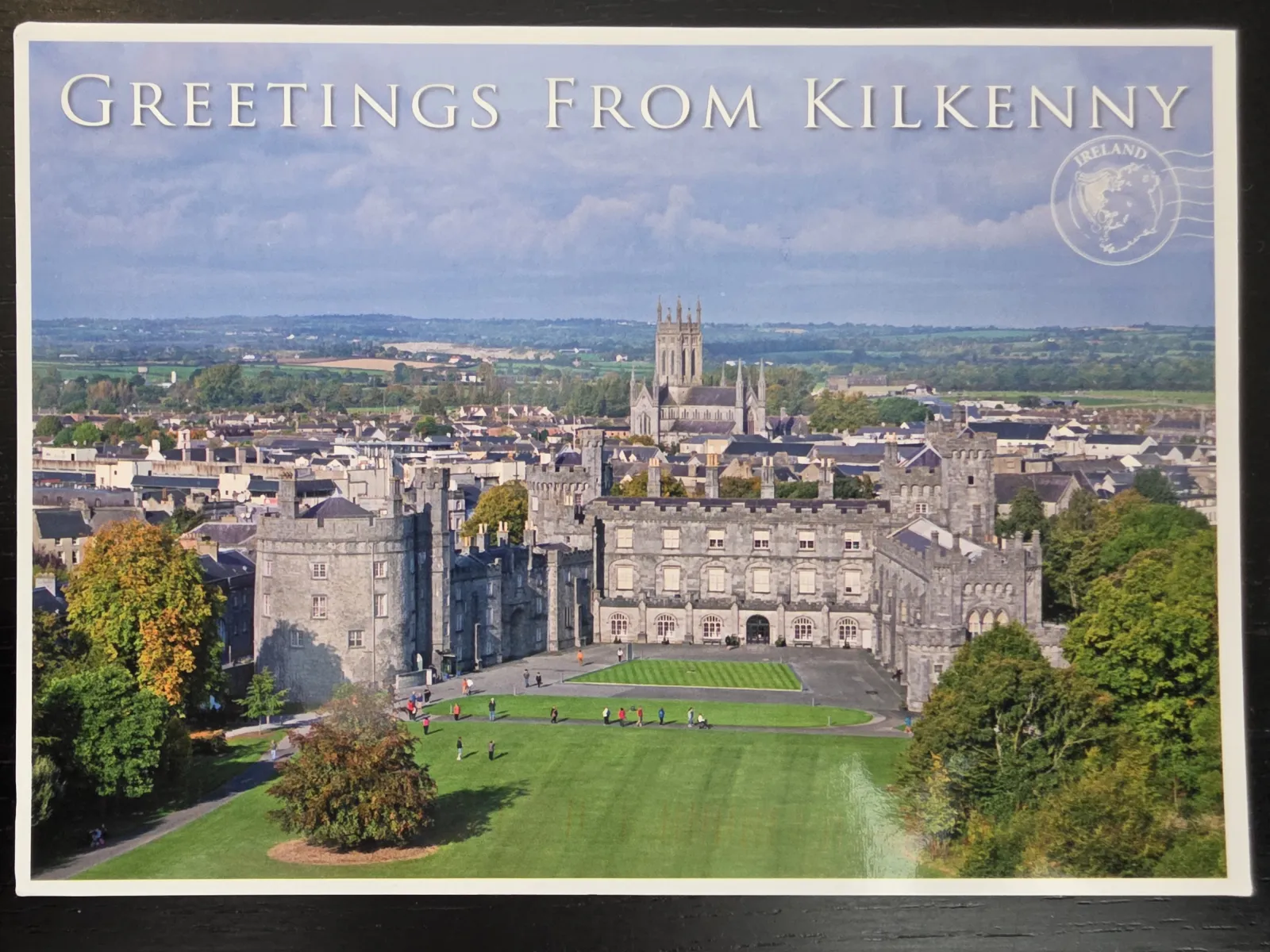 Eine Postkarte mit der Aufschrift „Greetings from Kilkenny“ zeigt das historische, graue Kilkenny Castle mit seinen grünen Rasenflächen vor der Kulisse der irischen Stadt und einer fernen Kathedrale.