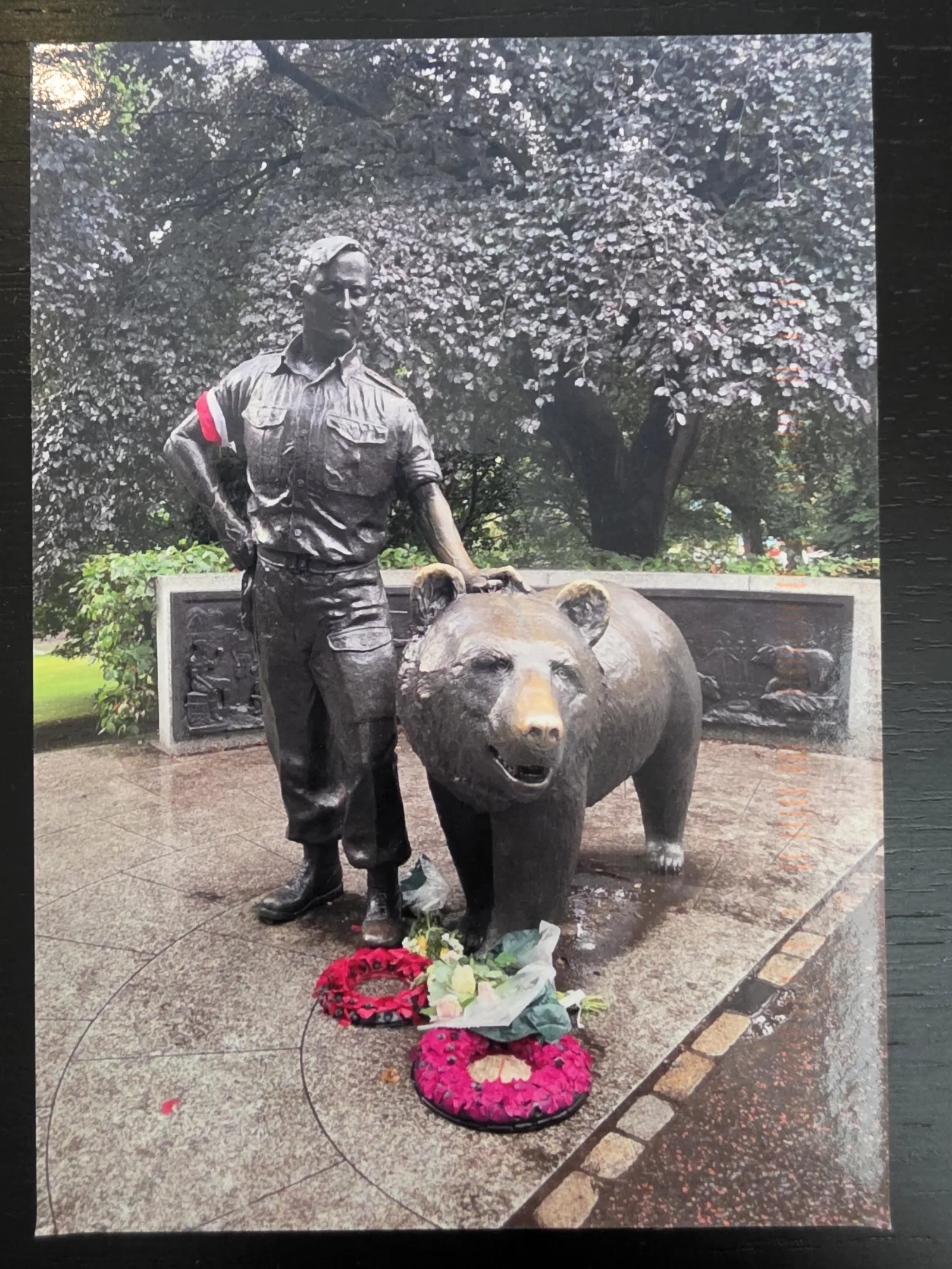 Eine Bronzestatue zeigt einen Soldaten in Uniform und einen großen Bären, vor denen zwei Blumenkränze auf dem Boden liegen.