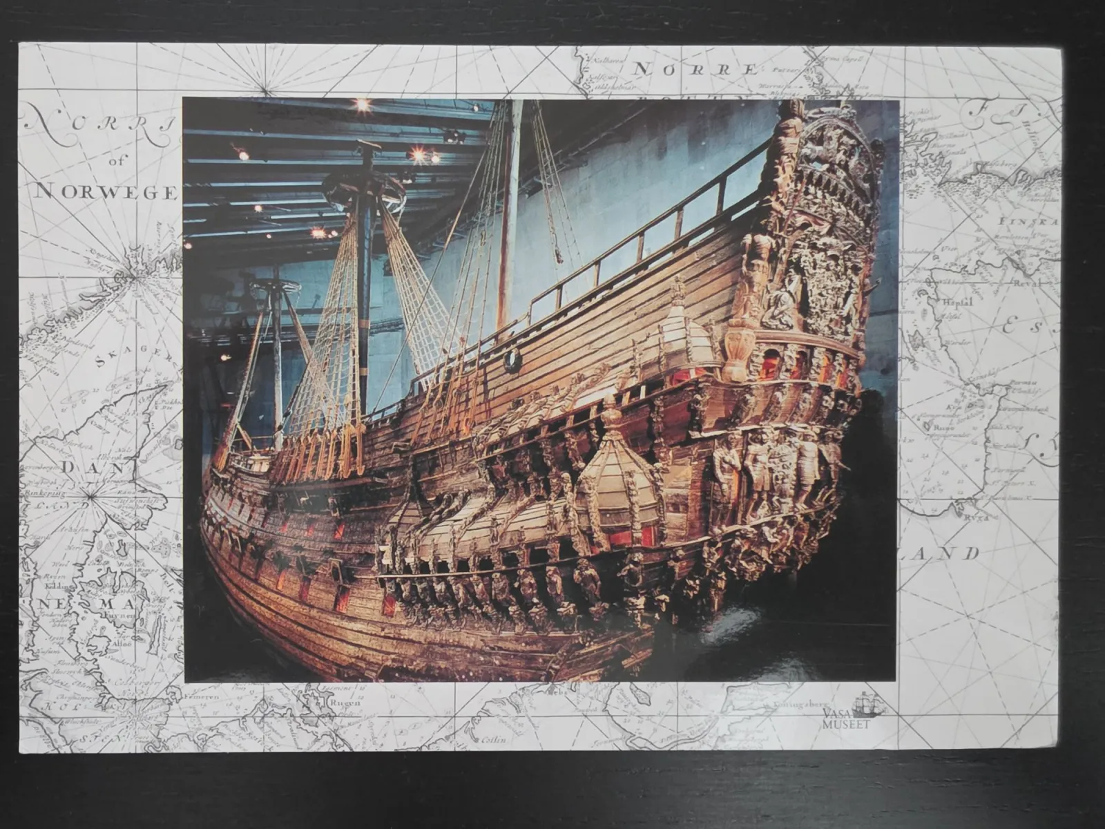 Eine Postkarte zeigt das reich verzierte Heck des historischen Holzschiffs Vasa vor dem Hintergrund einer alten Seekarte.