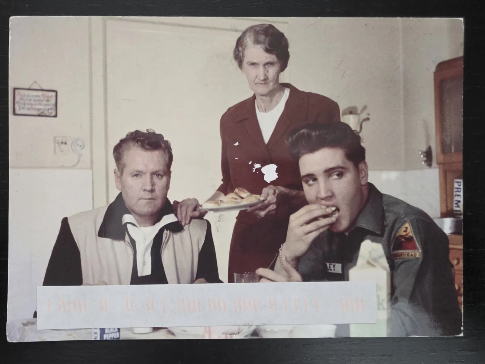 Ein historisches Foto zeigt den jungen Elvis Presley in Militäruniform beim Essen an einem Tisch, neben einem sitzenden Mann und einer älteren Frau, die Gebäck serviert.