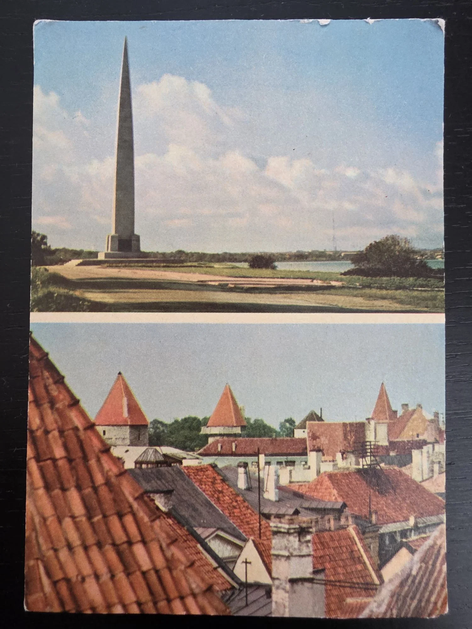 Eine zweigeteilte Postkarte zeigt oben einen hohen Obelisken in einer Küstenlandschaft und unten den Blick über die roten Ziegeldächer und mittelalterlichen Türme einer Altstadt.