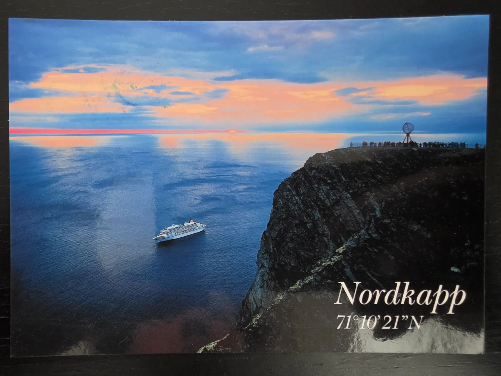 Das Bild zeigt das steile Kliff des Nordkapps mit dem Globus-Monument bei Sonnenuntergang, während ein Kreuzfahrtschiff auf dem tiefblauen Meer fährt.