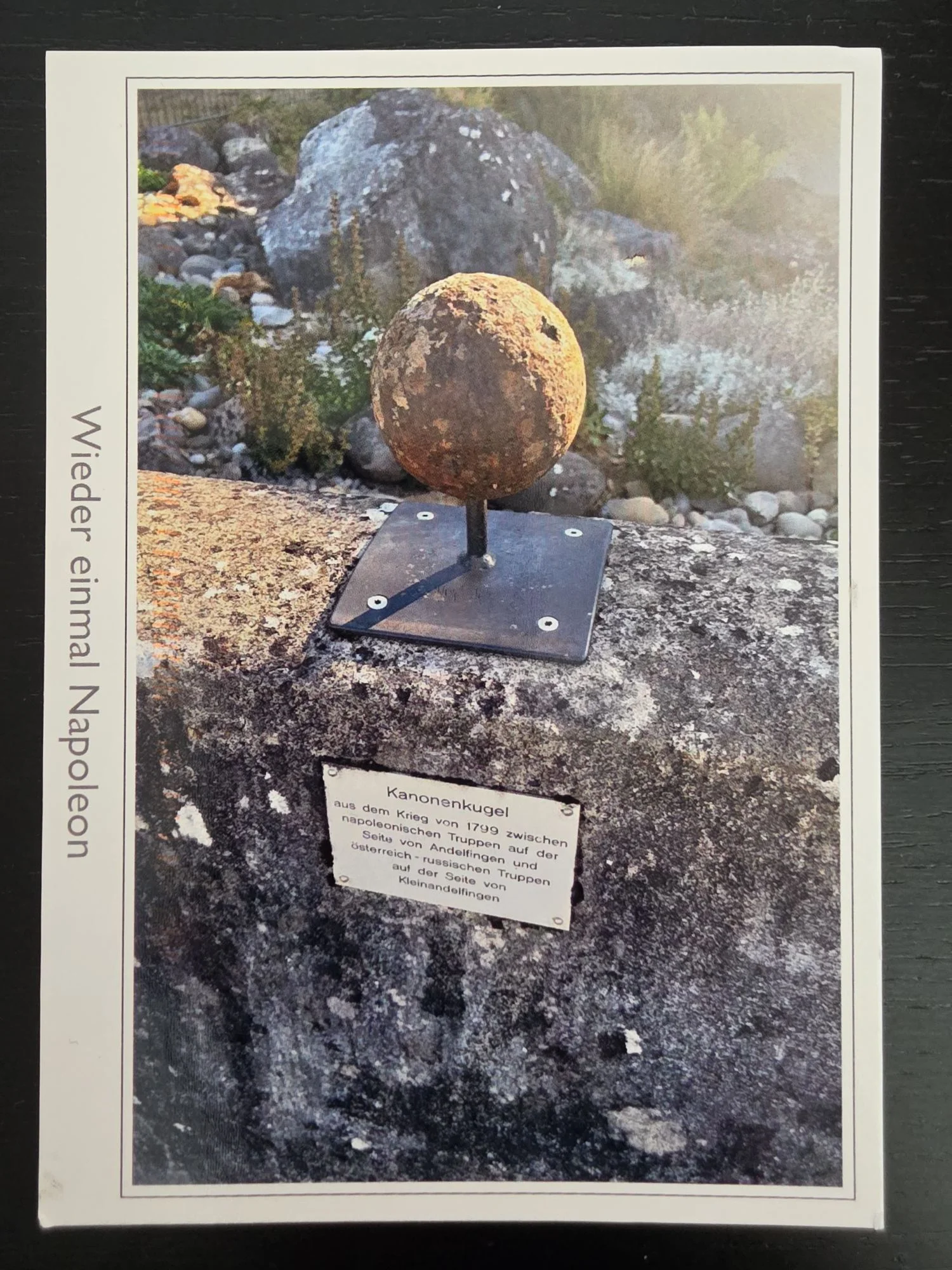 Eine auf einer Steinmauer montierte, rostige Kanonenkugel mit einer Informationstafel darunter, die an den Krieg von 1799 erinnert.