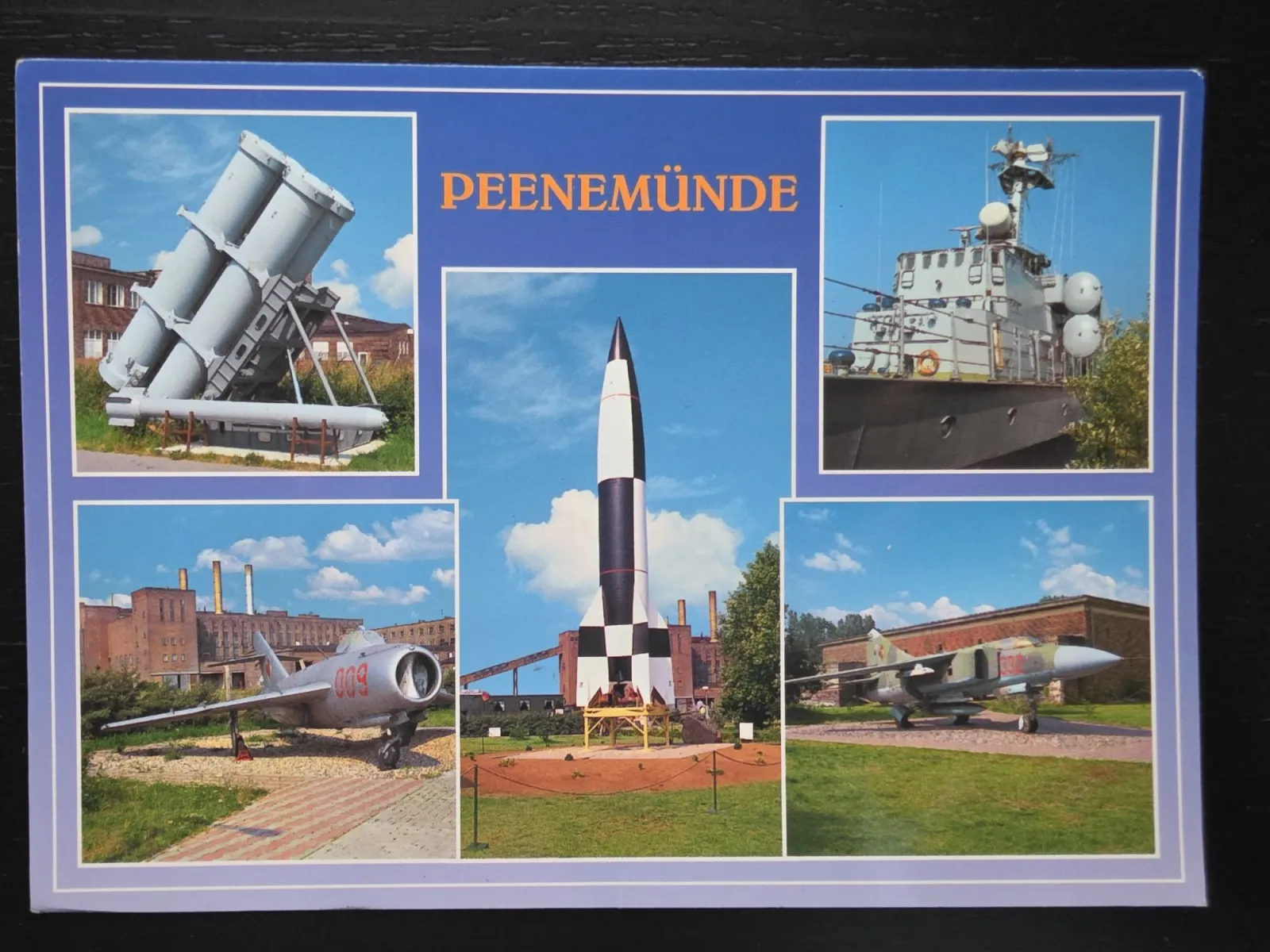 Eine Postkarte aus Peenemünde zeigt eine Collage historischer Militärexponate mit einer schwarz-weiß karierten V2-Rakete im Zentrum, umgeben von zwei Kampfjets, einem Kriegsschiff und einer Abschussrampe.