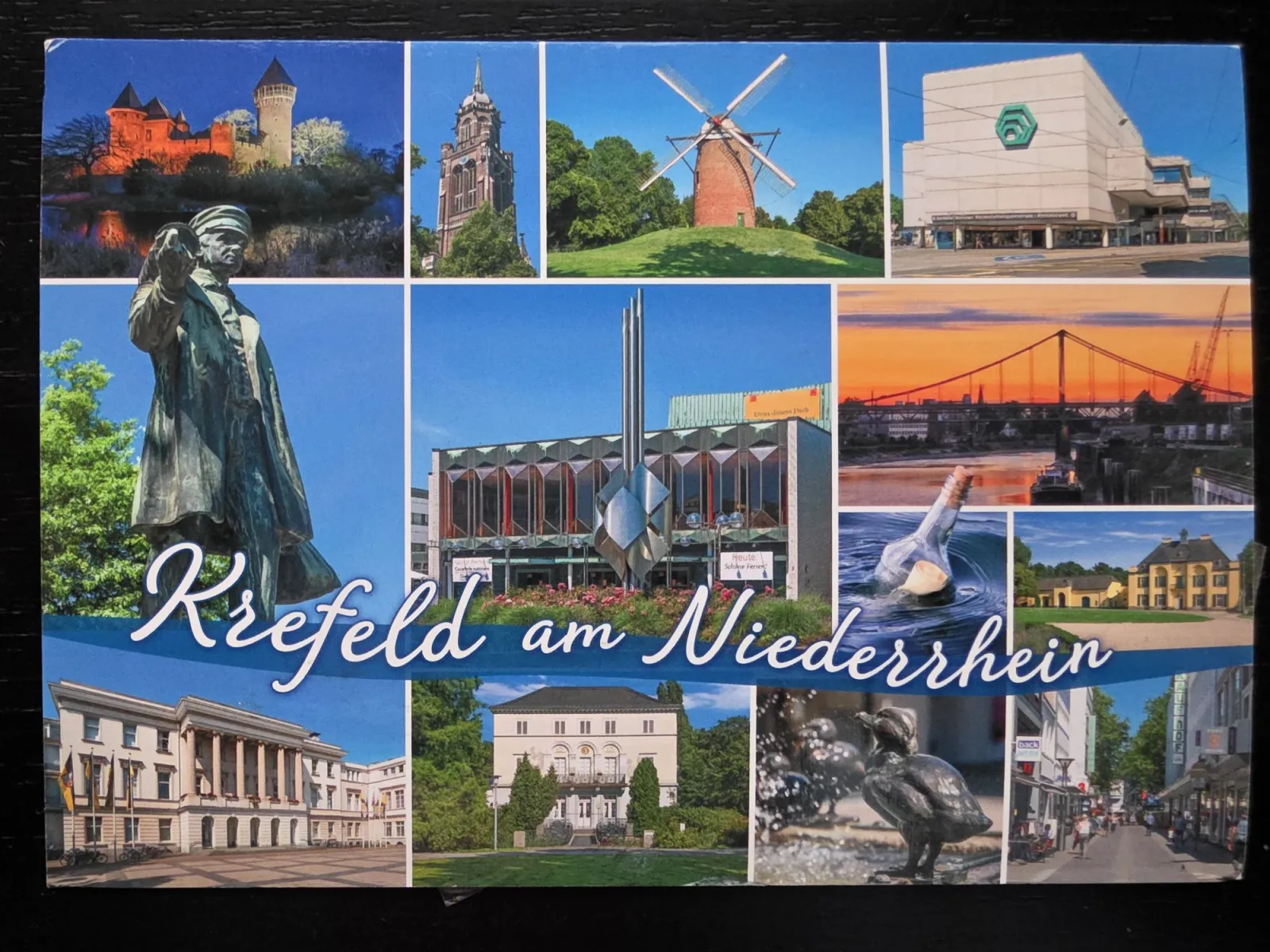 Eine Postkarten-Collage zeigt verschiedene Sehenswürdigkeiten der Stadt Krefeld, wie historische Gebäude, eine Windmühle, eine Brücke bei Sonnenuntergang und Statuen, überspannt von dem geschwungenen Schriftzug „Krefeld am Niederrhein“.