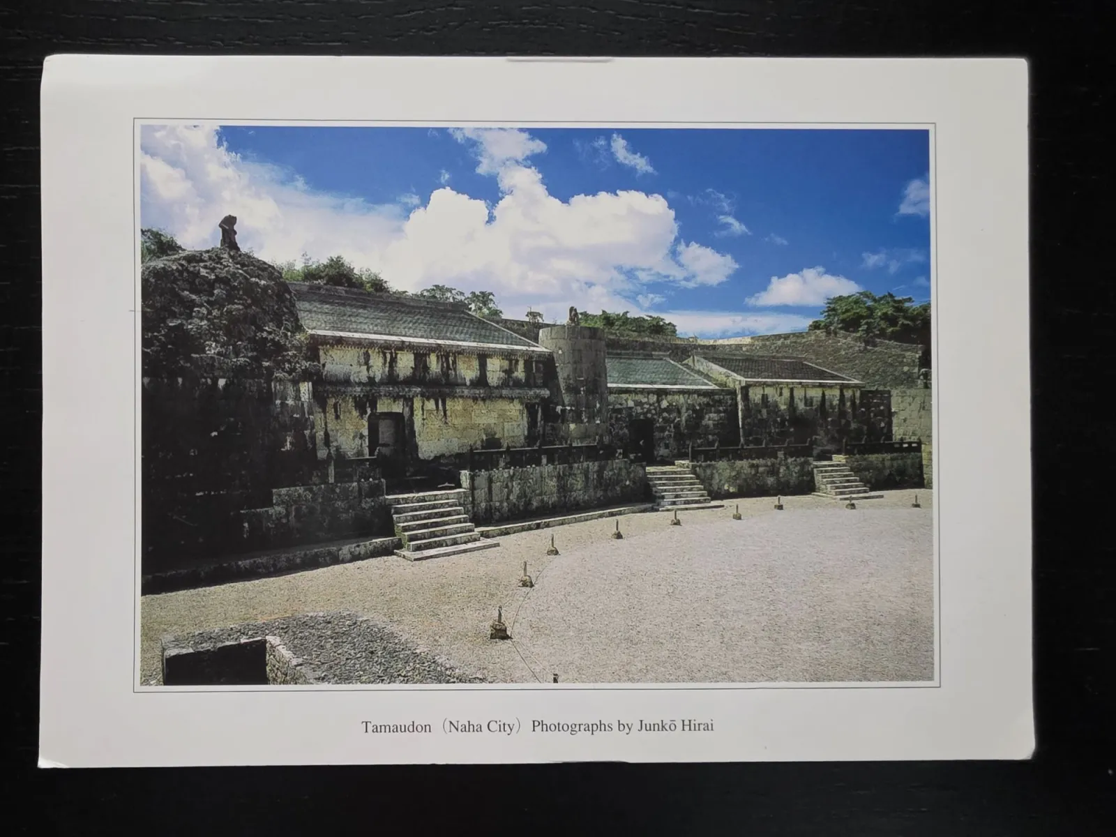 Eine Fotografie des historischen Steinmausoleums Tamaudon in Naha mit seinen alten Steingebäuden vor einem weiten Kiesplatz unter blauem, bewölktem Himmel.
