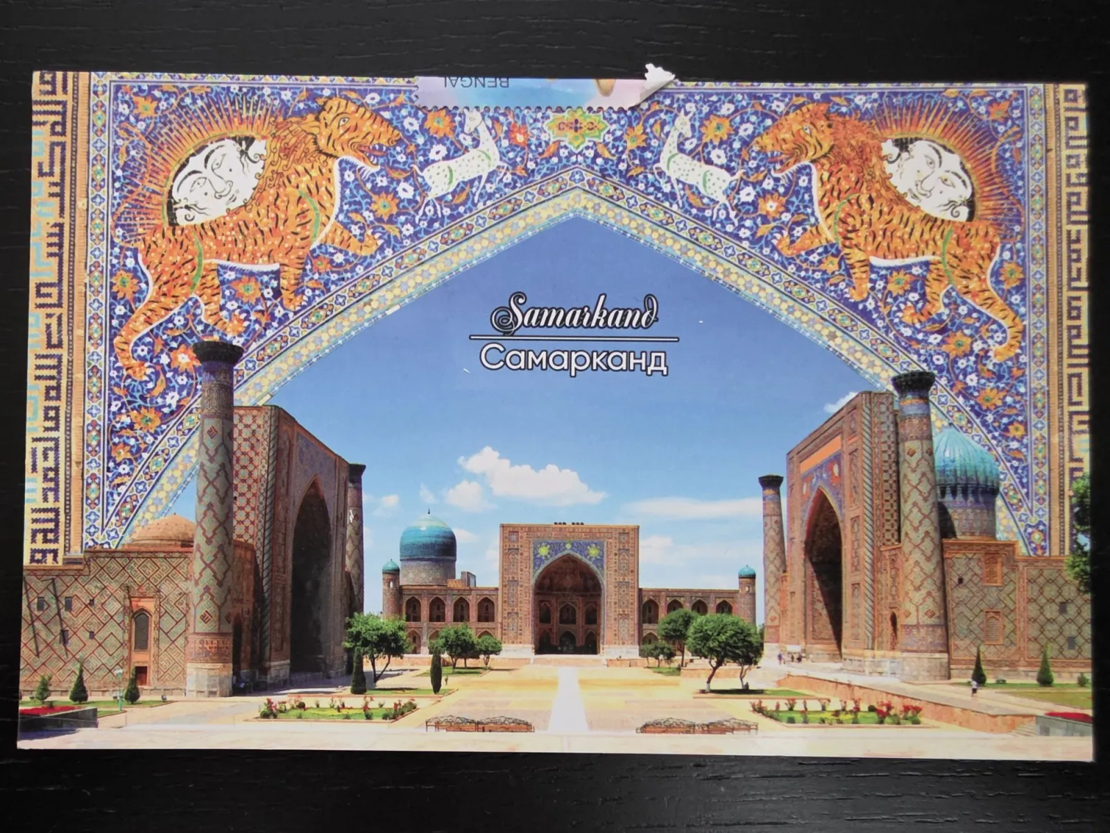 Eine Postkarte zeigt den historischen Registan-Platz in Samarkand, eingerahmt von einem kunstvollen, blauen Mosaikbogen mit Tigern und Sonnenmotiven.