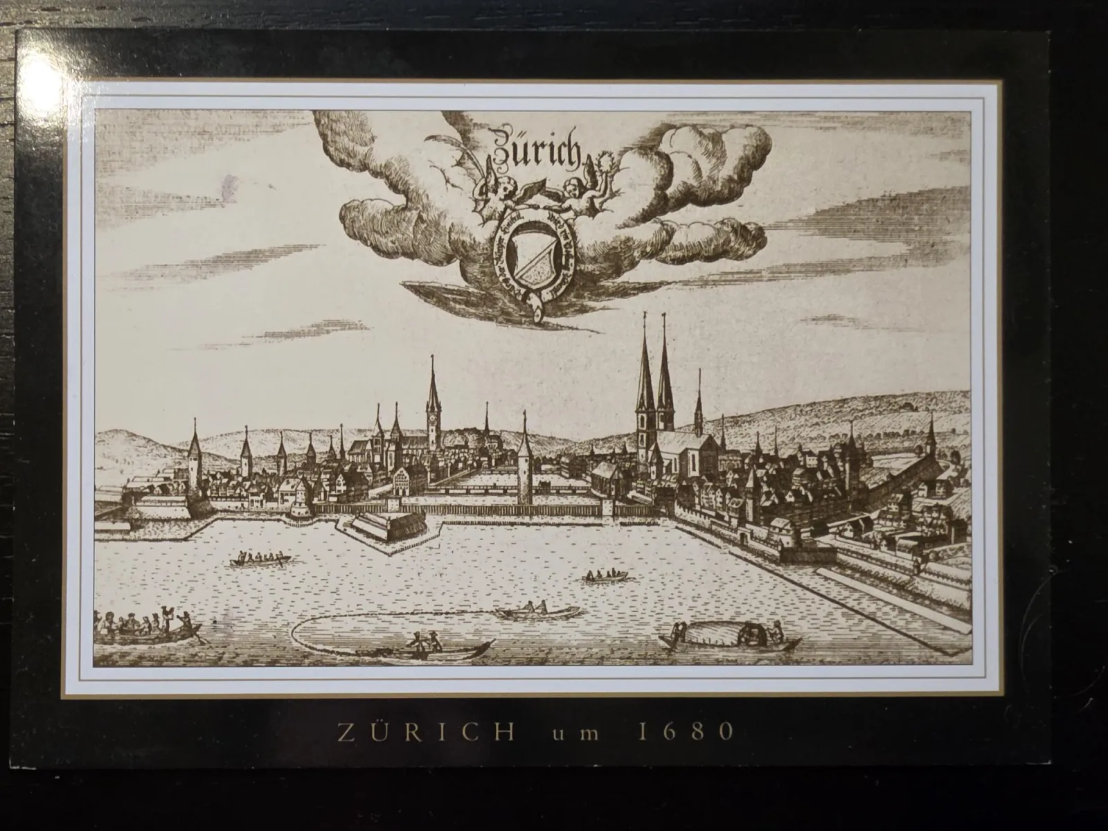 Ein historischer Schwarz-Weiß-Kupferstich zeigt die Panoramaansicht der Stadt Zürich um 1680 vom See aus mit mehreren Booten im Vordergrund und einem von Engeln gehaltenen Wappen im Wolkenhimmel.