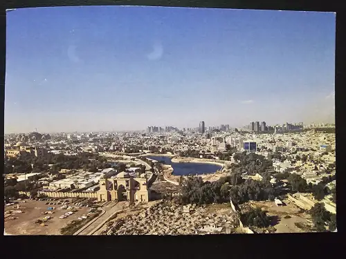 Ein Panoramabild zeigt eine weite Stadtansicht unter blauem Himmel, mit einem großen, hellen Tor im Vordergrund, einem Fluss und Hochhäusern am Horizont.