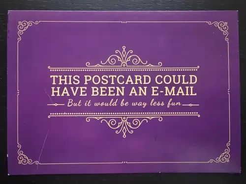 Eine violette Postkarte mit goldenem Zierrahmen und dem englischen Text: „THIS POSTCARD COULD HAVE BEEN AN E-MAIL – But it would be way less fun –“.