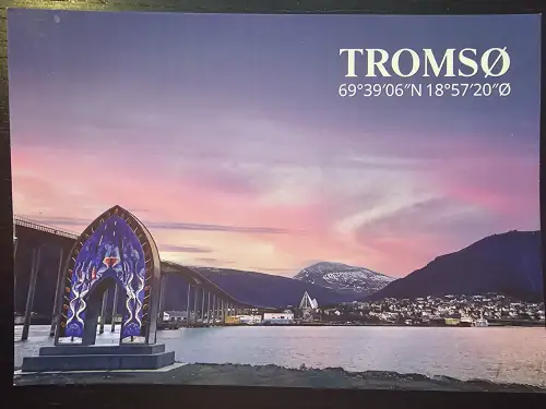 Tromsø bei Sonnenuntergang, mit einem markanten blauen Bogen im Vordergrund, der Stadt am Wasser und Bergen unter einem rosa-lila Himmel.