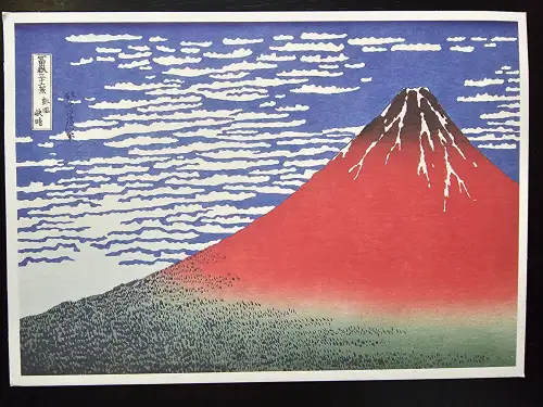 Der japanische Holzschnitt zeigt den rotbraunen Berg Fuji mit weißen Schneeflecken, einem blauen Himmel mit weißen Wolken und grünen Wäldern am Fuße.