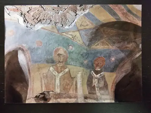 Ein sehr altes, stark beschädigtes Fresko zeigt zwei religiöse Figuren mit Heiligenscheinen unter geometrischen Mustern, wobei oben Mauerwerk freiliegt.