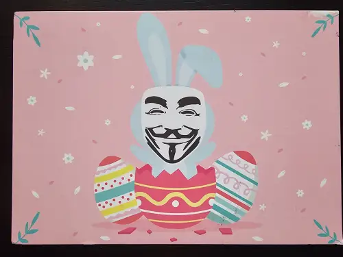 Auf rosafarbenem Hintergrund schlüpft ein Osterhase mit Guy-Fawkes-Maske aus einem zerbrochenen Osterei, flankiert von zwei weiteren bunten Eiern und kleinen Blumen.