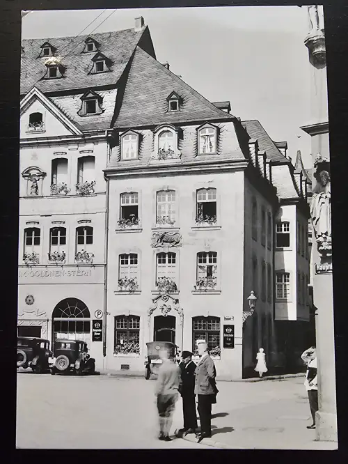 Ein Schwarz-Weiß-Foto zeigt eine belebte historische Straße mit mehrstöckigen Gebäuden, geparkten Oldtimern und mehreren Personen.